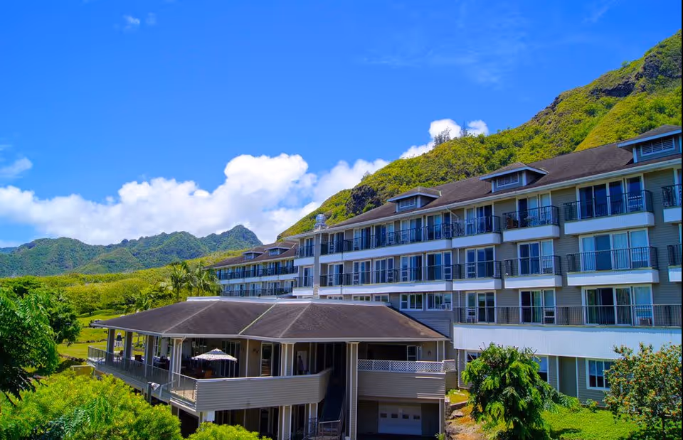 A multi-story senior living building with balconies beside green hills and a blue sky.
