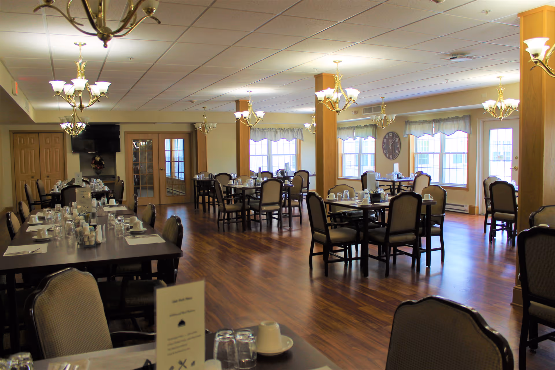 Spacious dining room with multiple set tables and chairs, chandeliers, wood floors, and large windows.