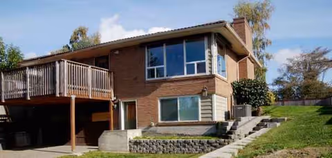 Two-story brick house exterior with a wooden deck, stairs, and green lawn.
