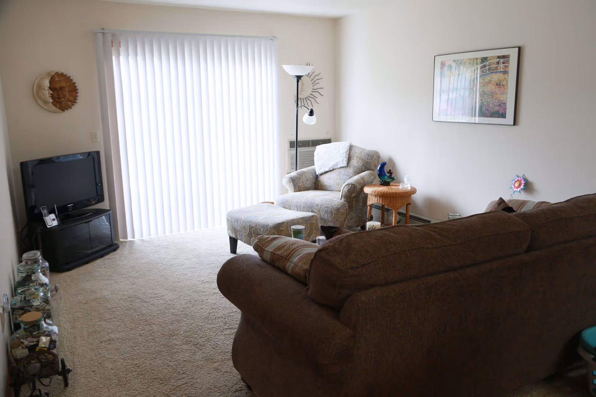 A cozy living room with a brown couch in the foreground and a patterned armchair with matching ottoman near a sliding glass door covered by vertical blinds. A small wicker side table with decorative items sits next to the armchair. A floor lamp stands behind the armchair, and a flat-screen TV is placed on a black stand in the corner. The walls are light-colored with a framed picture and sun and moon wall decorations.