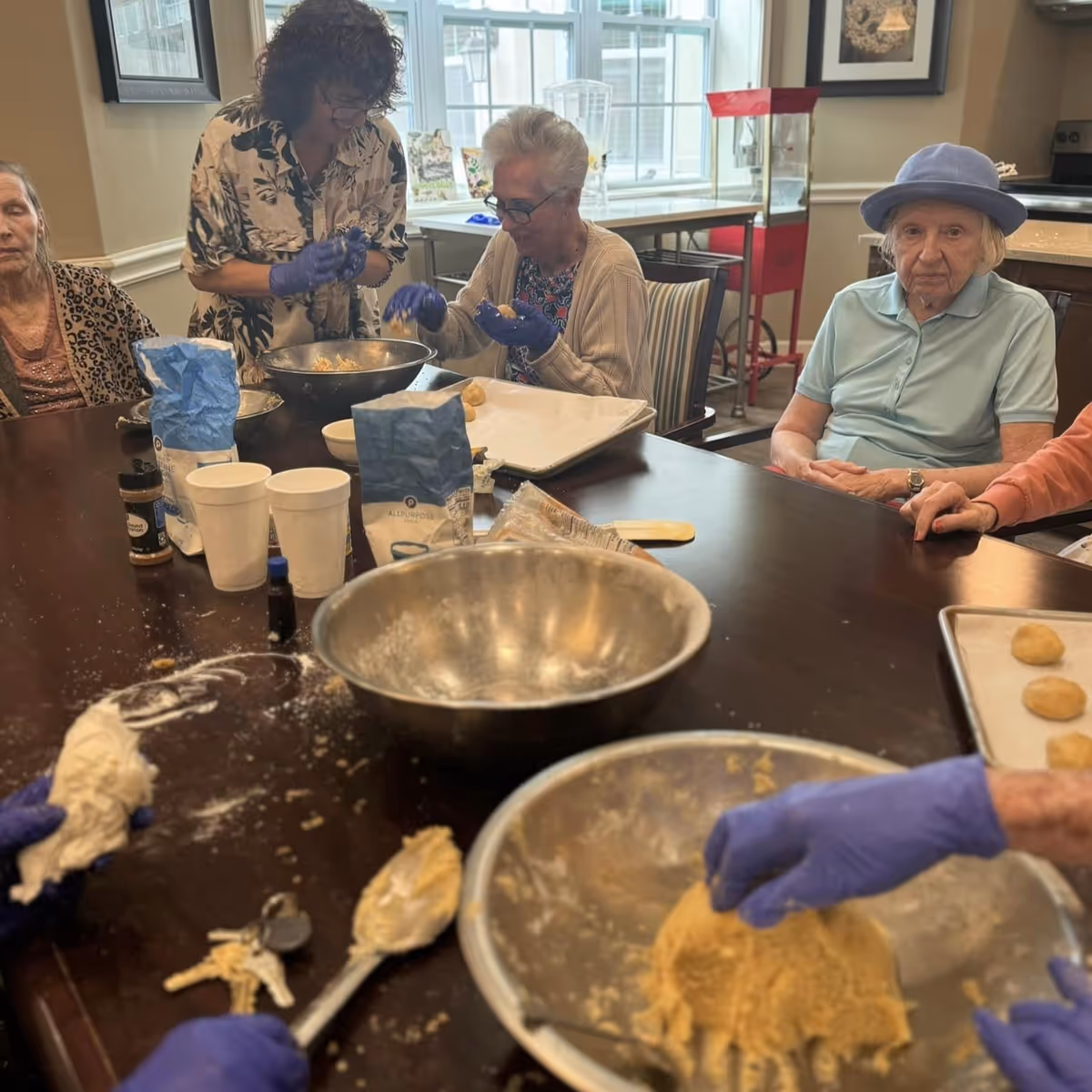 Elderly residents wearing gloves sit around a table mixing and forming cookie dough during a group activity.