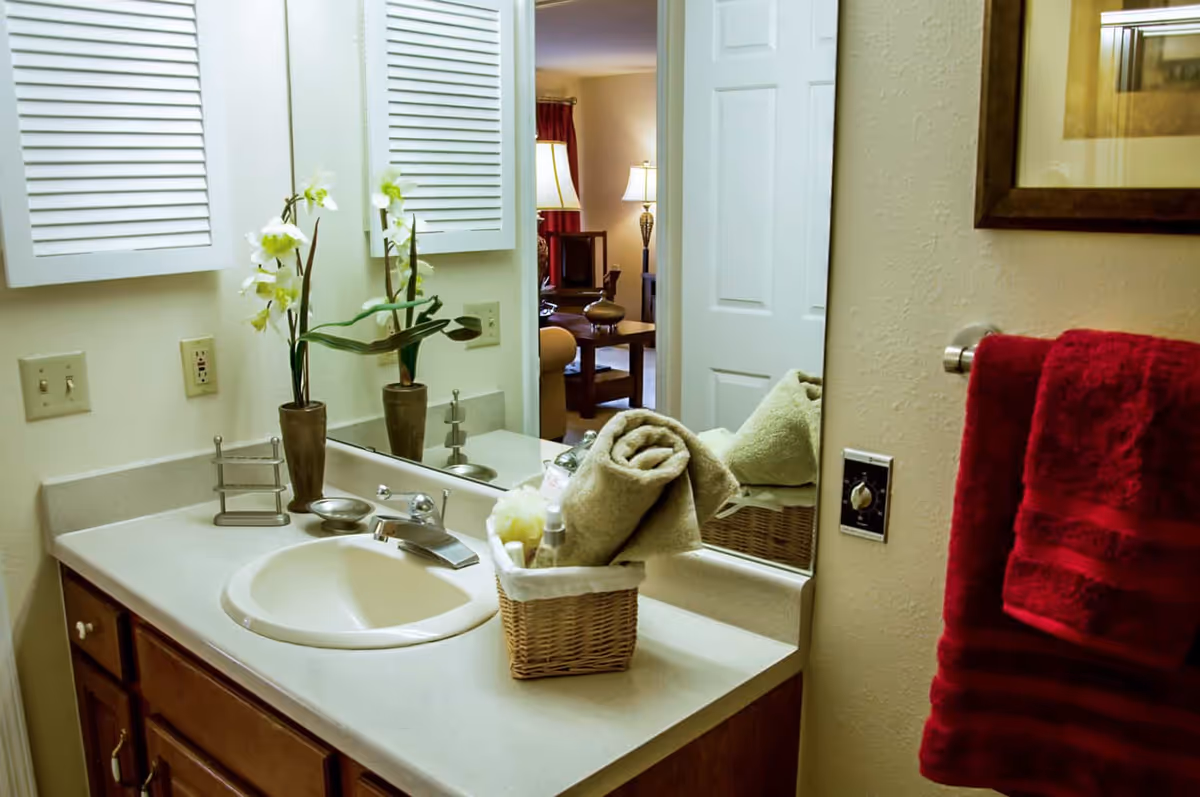 Bathroom sink area with a beige countertop, a round sink, and a silver faucet. A wicker basket on the counter holds rolled towels and toiletries. A vase with white flowers is placed near the mirror. A red towel hangs on a towel rack on the wall. In the background, an open door reveals a living room with a lamp and furniture.