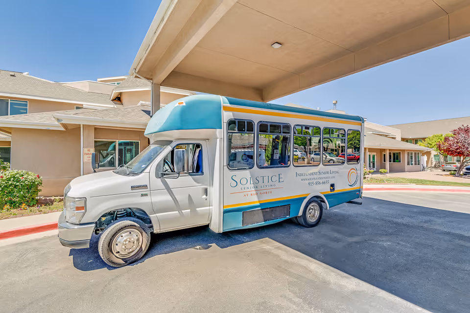A white and teal shuttle bus parked under a covered driveway at Solstice Senior Living at Rio Norte facility. The bus has the facility's name and contact information on its side. The building and some landscaping are visible in the background under a clear blue sky.