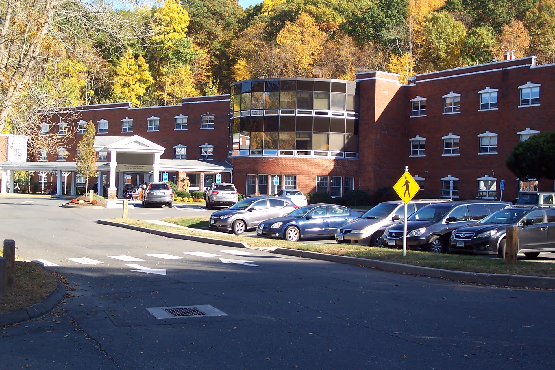Front exterior of a brick health and rehabilitation building with a covered entrance, parked cars, and autumn trees behind.
