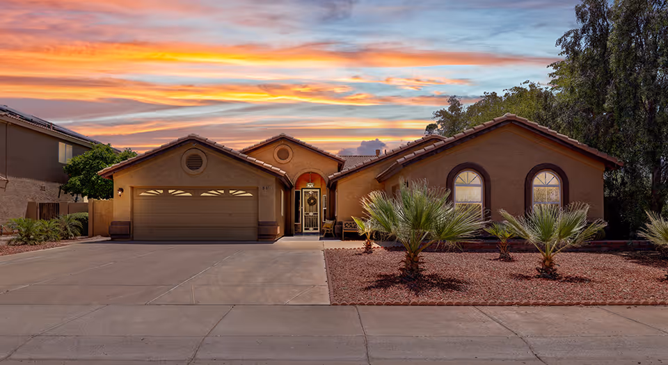 Single-story residential house with a two-car garage, arched windows, and a front entrance with a small porch, set against a colorful sunset sky. The front yard features desert landscaping with small palm plants and red gravel.