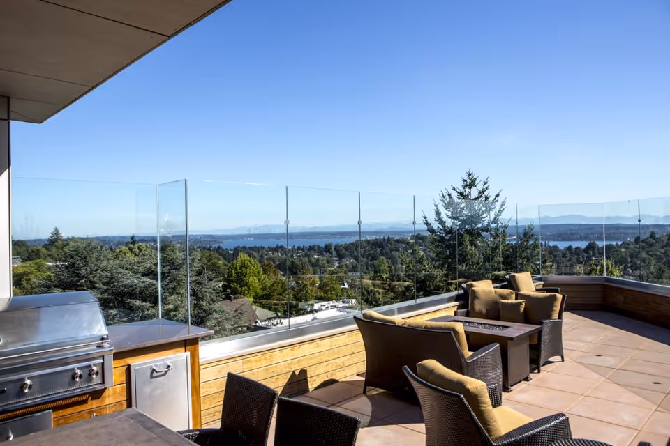Outdoor patio area with cushioned wicker chairs and a fire pit table, a stainless steel grill, glass railing, and a scenic view of trees, water, and distant mountains under a clear blue sky.