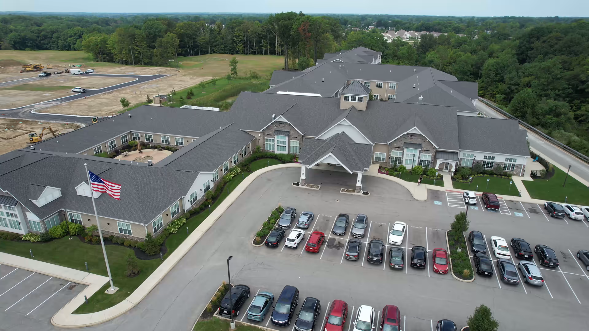 Aerial view of Vitalia Senior Residences at Strongsville showing a large, single-story building with a gray roof and beige exterior walls. The building has a covered entrance with a driveway and a parking lot filled with cars. Surrounding the building are green lawns, trees, and a wooded area in the background.