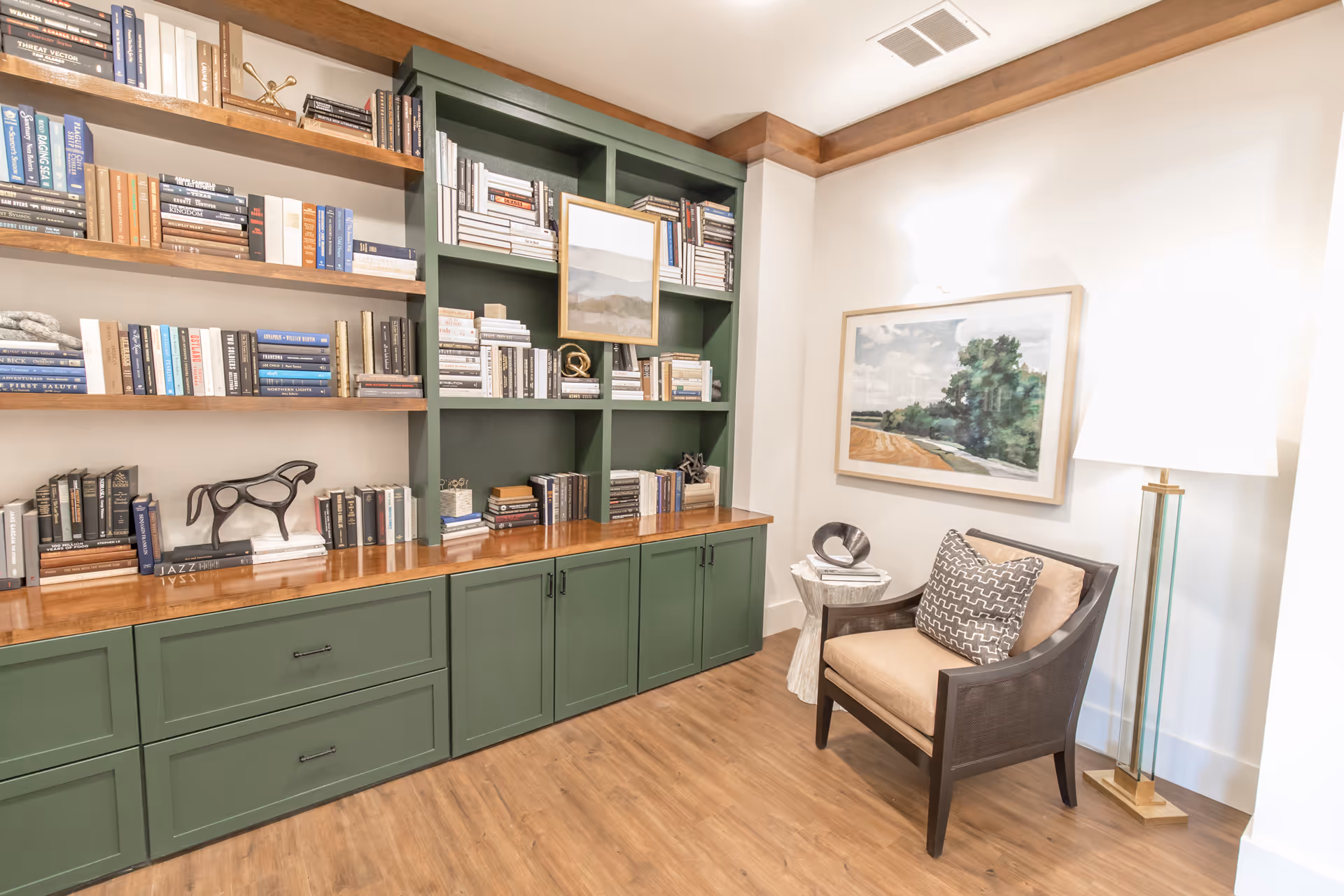 A cozy reading nook featuring green built-in bookshelves filled with books and decorative items, a comfortable armchair with a patterned pillow, a small side table with a sculpture, a floor lamp, and a framed landscape painting on the wall.