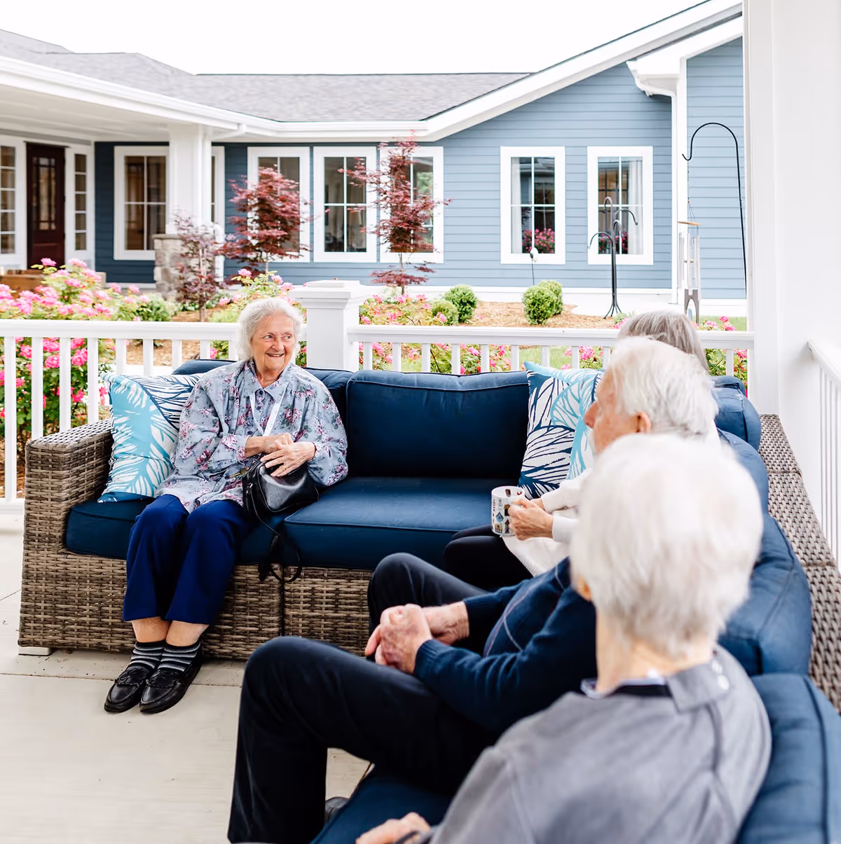A group of elderly people sitting and chatting on a covered outdoor patio with blue cushioned wicker furniture. The background shows a well-maintained garden with flowers and shrubs, and a blue building with white trim windows.