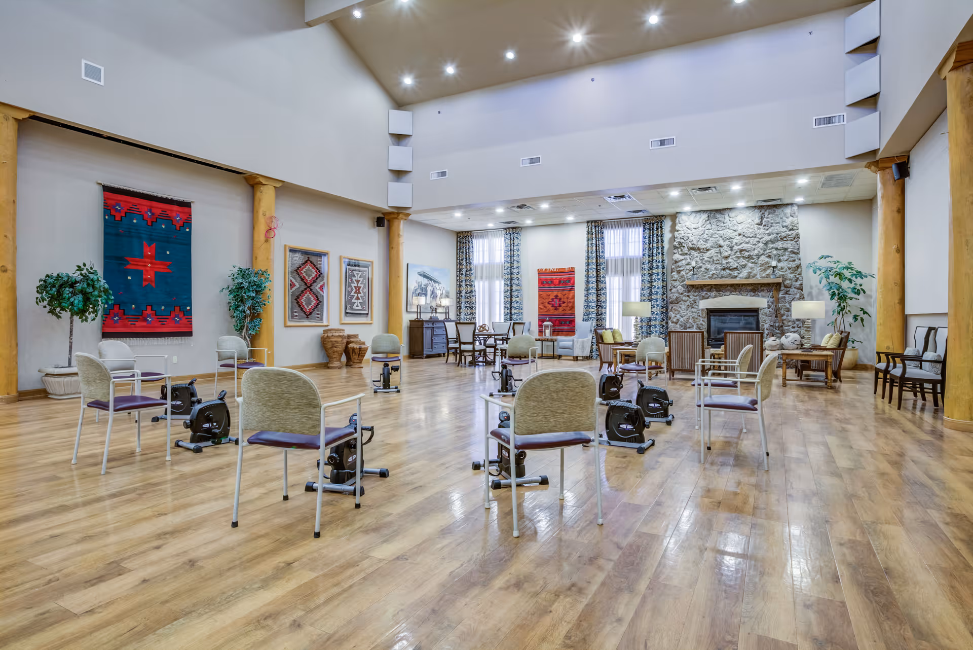 A spacious senior living community room with wooden floors, several chairs arranged in a circle each paired with a small exercise bike, large windows with patterned curtains, colorful wall hangings, potted plants, and a stone fireplace in the background.