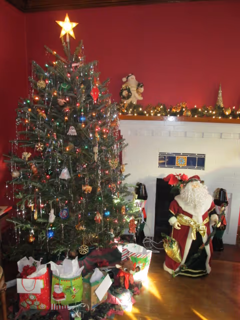 Decorated Christmas tree with lights and wrapped presents beside a fireplace and a large Santa figure.