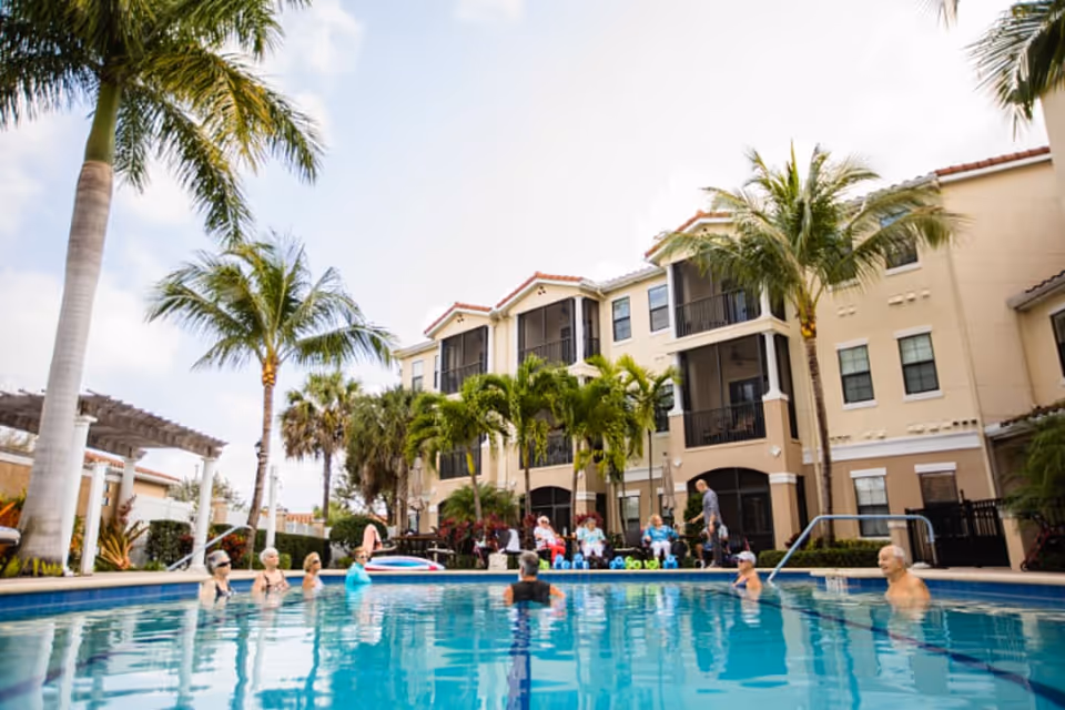A group of elderly people enjoying a swim in an outdoor pool surrounded by palm trees, with a multi-story residential building in the background under a partly cloudy sky.