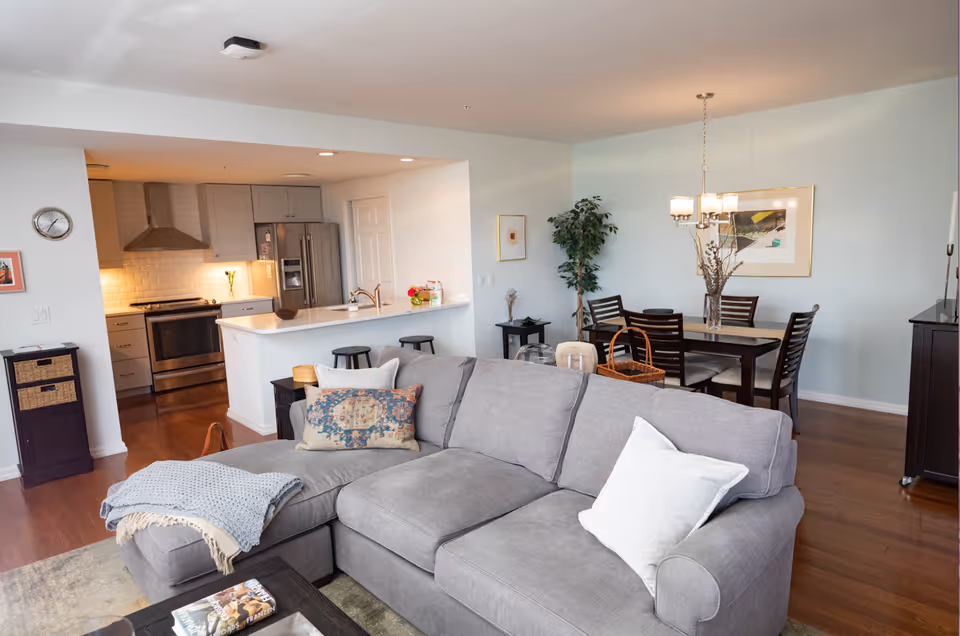 Open concept living area featuring a gray sectional sofa with pillows and a throw blanket, a coffee table with magazines, a dining table with six chairs and a chandelier above, and a modern kitchen with stainless steel appliances and a breakfast bar with stools.