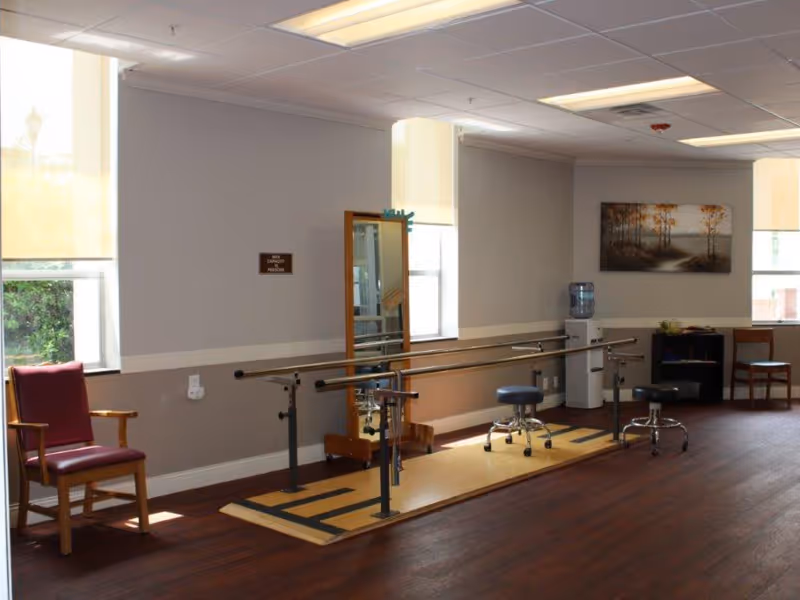 Interior therapy room with parallel walking bars, rolling stools, chairs, a mirror and a water cooler.