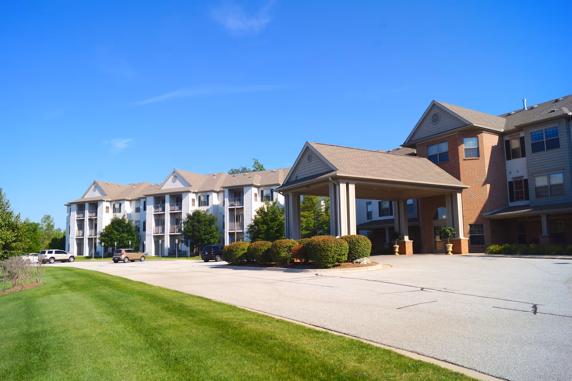 Exterior view of the Village At Arborwood of Mishawaka building with a covered entrance, parking area, and green lawn under a clear blue sky.