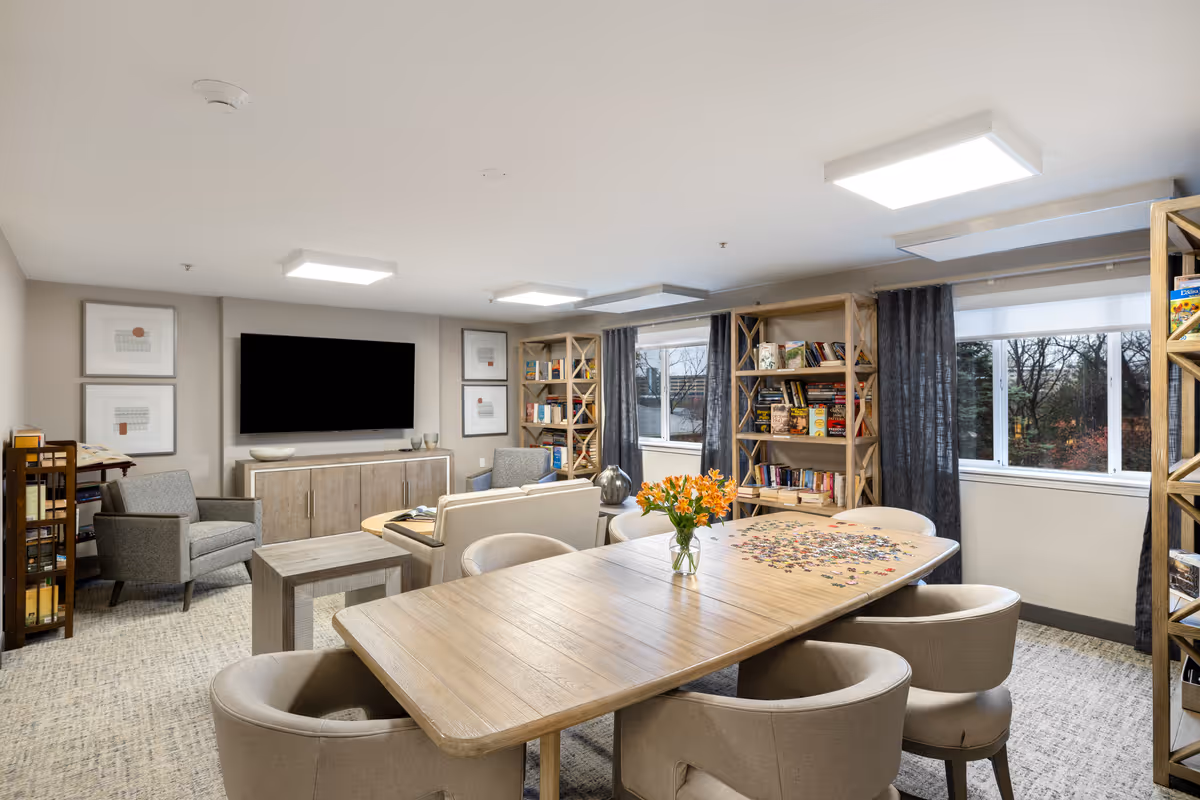 A well-lit common room with a large wooden table surrounded by six cushioned chairs. On the table, there is a vase with orange flowers and a partially completed jigsaw puzzle. The room has two large windows with dark curtains, wooden bookshelves filled with books and games, a flat-screen TV mounted on the wall, and comfortable armchairs arranged around a small coffee table.