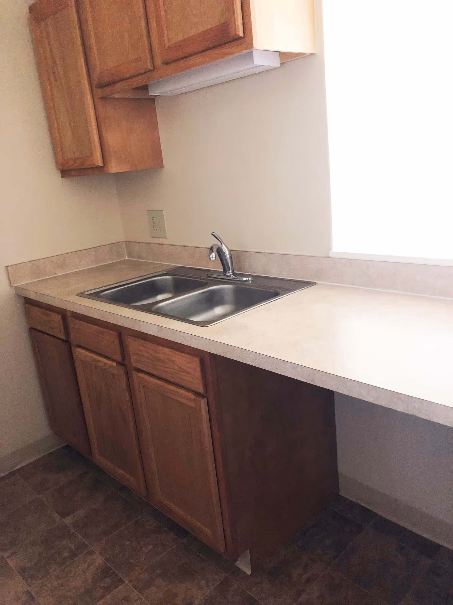 Small kitchen countertop with a double stainless steel sink, wooden cabinets and a window.