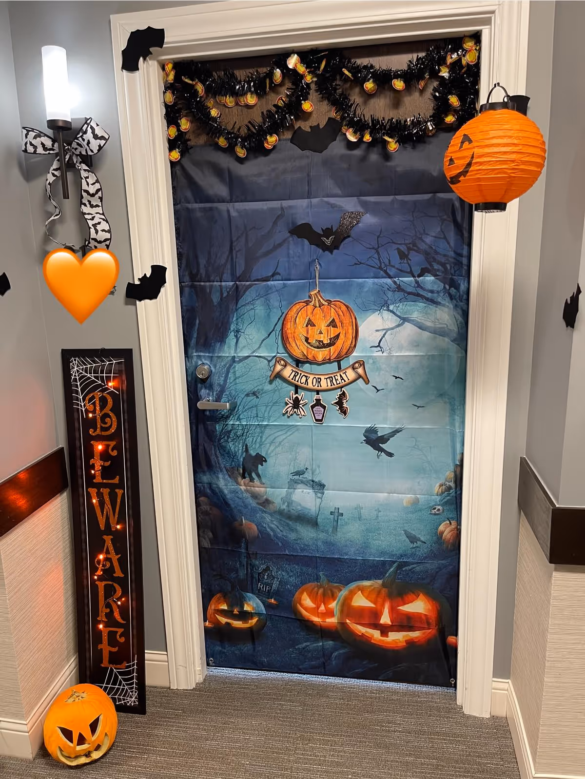 Hallway apartment door decorated for Halloween with a spooky door cover, jack-o'-lanterns, bats, garland and a "Trick or Treat" sign.