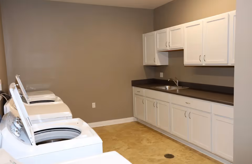 A clean laundry room with several top-loading washing machines along the left wall and white cabinets and a sink along the right.
