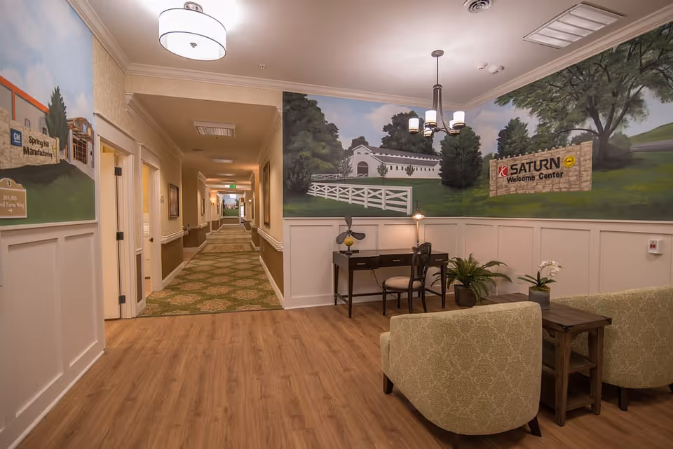 A well-lit senior living facility interior hallway with seating area, desk, and a large mural on the wall.