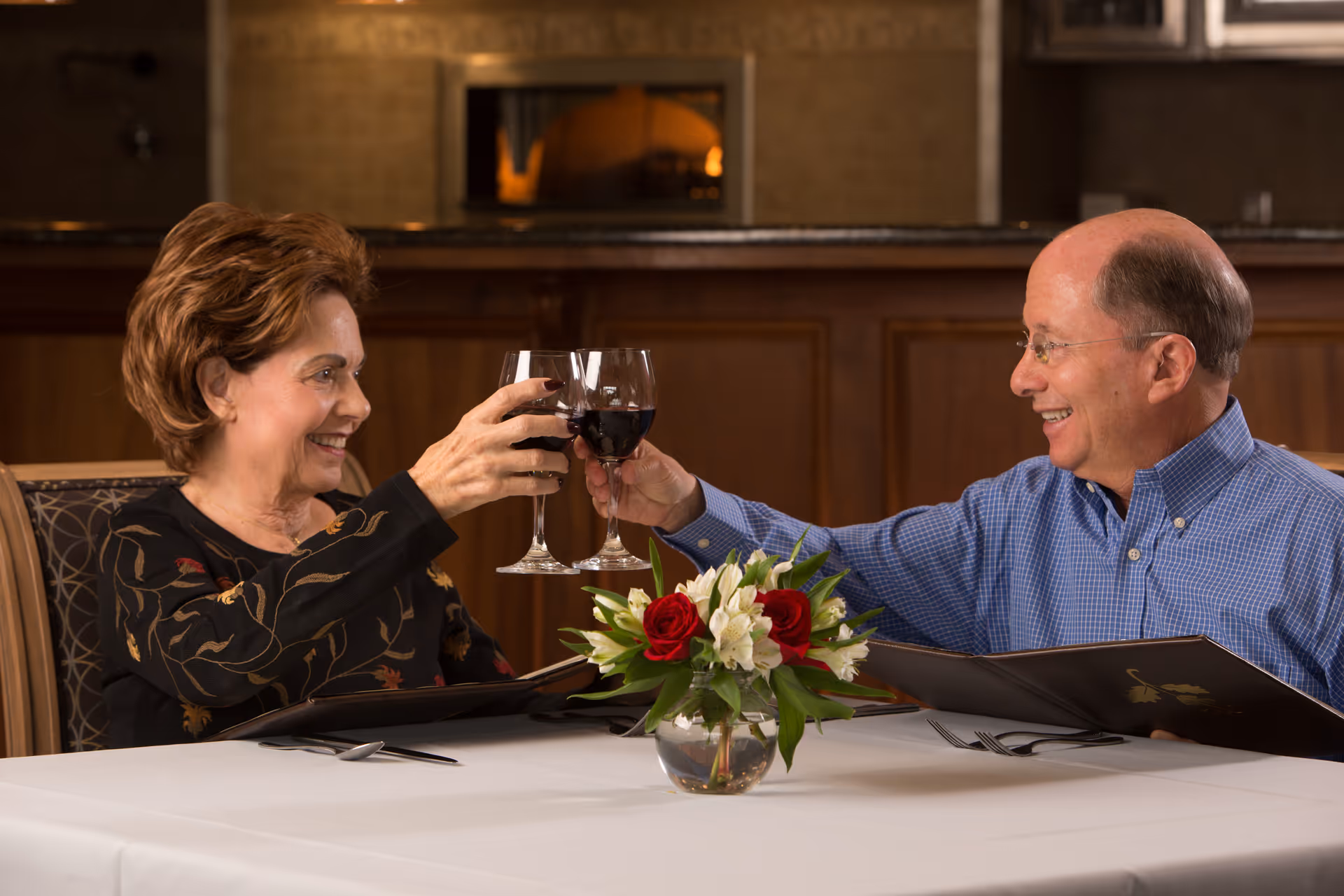 An elderly woman and man sitting at a dining table in a restaurant, smiling and clinking their wine glasses filled with red wine. There is a small vase with red and white flowers on the table, and both are holding menus.