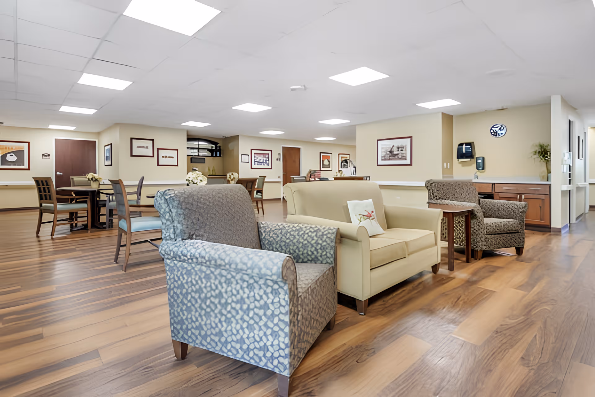 A spacious and well-lit common area in a senior living facility featuring comfortable armchairs and a sofa arranged on a wooden floor. Several tables and chairs are visible in the background along with framed artwork on beige walls. The ceiling has recessed lighting panels, and there is a countertop with a paper towel dispenser and hand sanitizer on the right side.