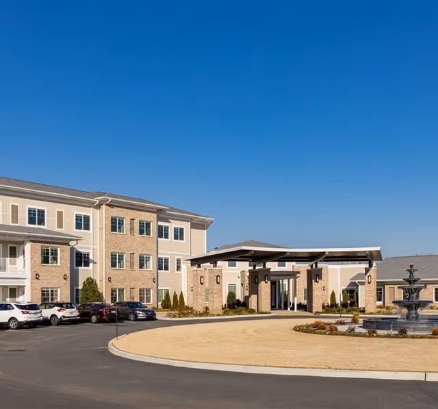 Front exterior of a multi-story senior living building with a covered entrance, circular driveway, parked cars, and a fountain under a clear blue sky.