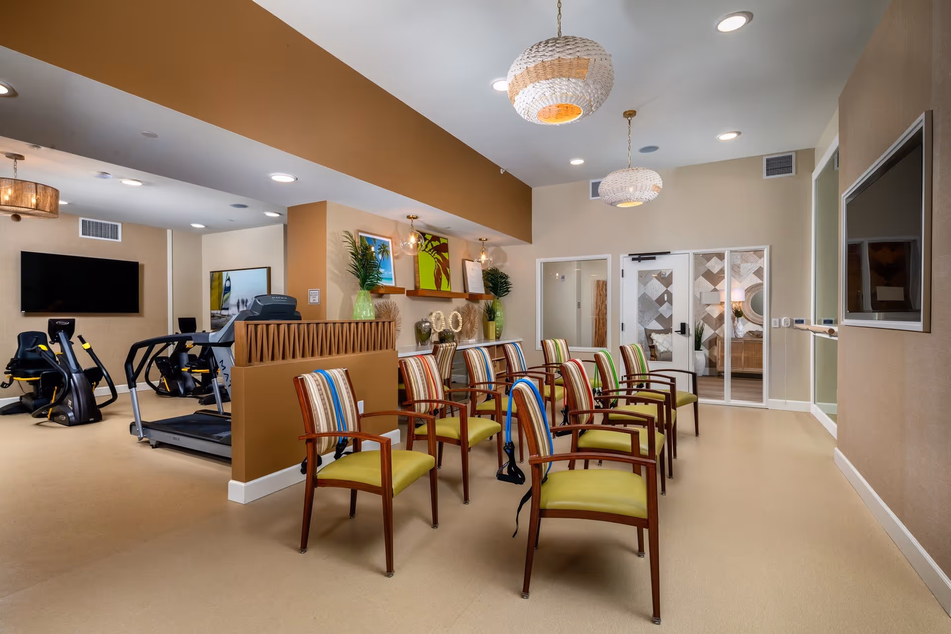 Bright communal interior with rows of colorful chairs facing a TV and nearby exercise equipment in a senior living activity room.
