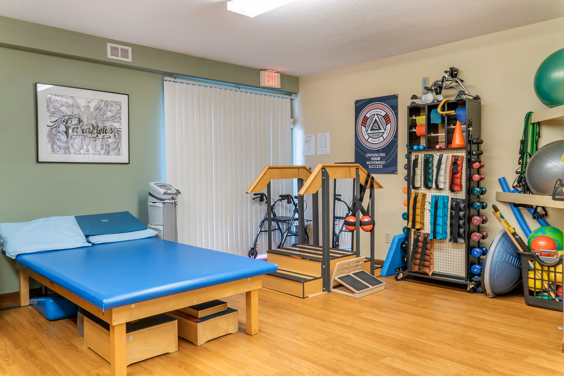 A physical therapy room with a blue padded treatment table, a set of wooden stairs with handrails for rehabilitation exercises, various exercise equipment including weights, resistance bands, and therapy balls, and a walker near a window with vertical blinds.