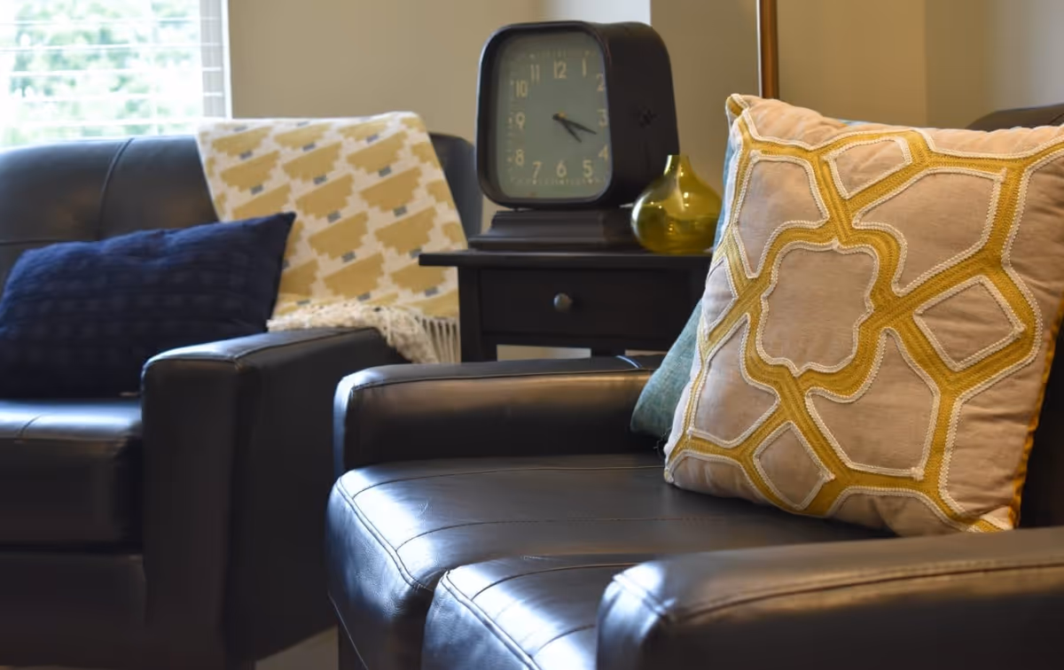 Close-up view of two black leather armchairs with decorative pillows, one with a beige and yellow patterned pillow and the other with a dark blue pillow and a beige and yellow throw blanket. A black side table between the chairs holds a vintage-style clock and a small yellow glass vase. A window with blinds is visible in the background.