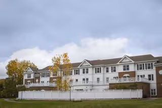 Exterior view of a multi-story senior living facility building with white siding and brick accents, surrounded by a white fence and green lawn, under a cloudy sky.