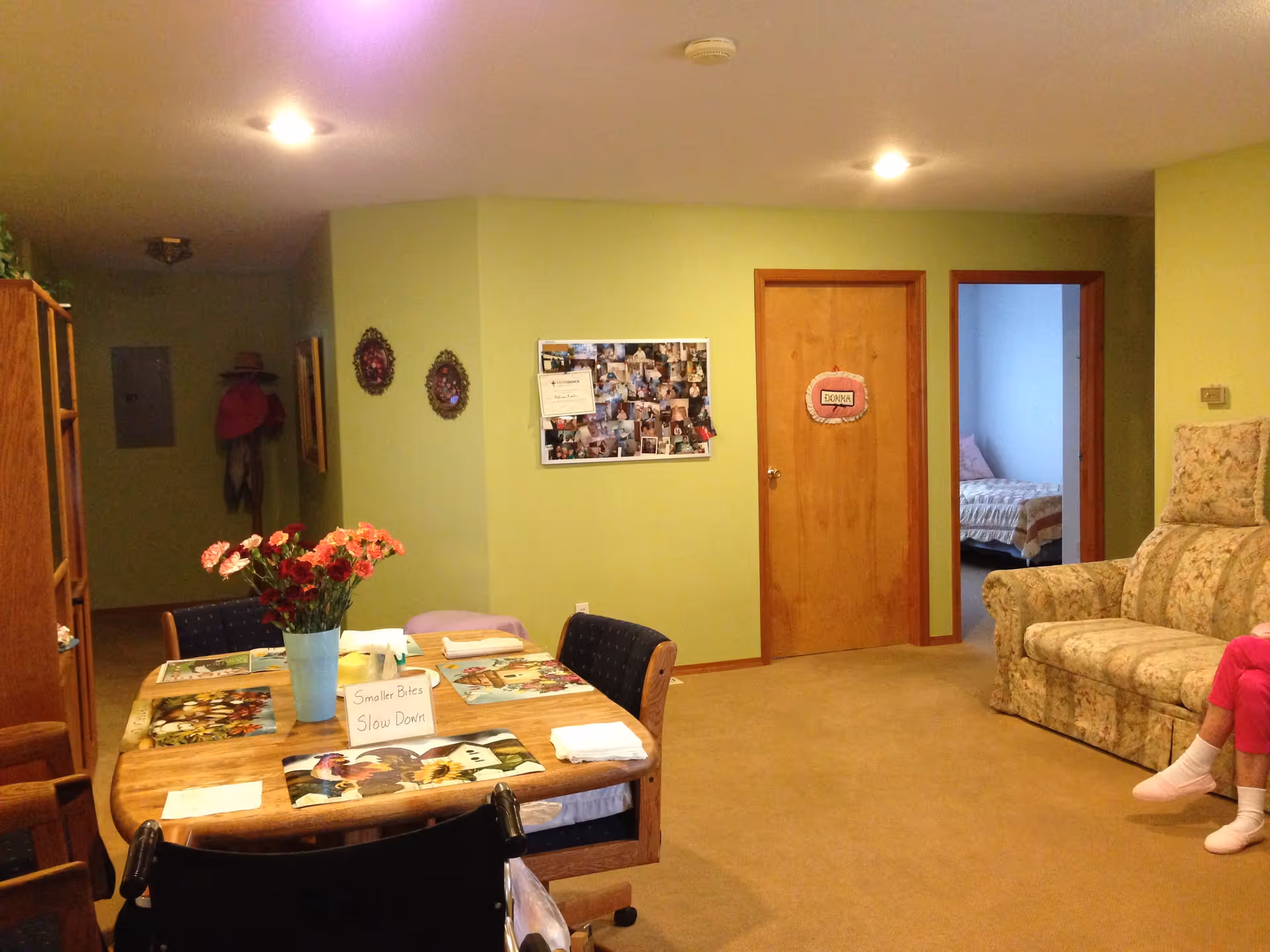 Interior of a senior living facility room with light green walls, a wooden dining table with floral placemats and a vase of flowers, a floral patterned couch, and a person sitting on the right side wearing pink pants and white socks. There are two wooden doors on the far wall, one with a sign that reads 'Donna', and a bulletin board with photos and papers. A bedroom with a bed is visible through an open doorway.