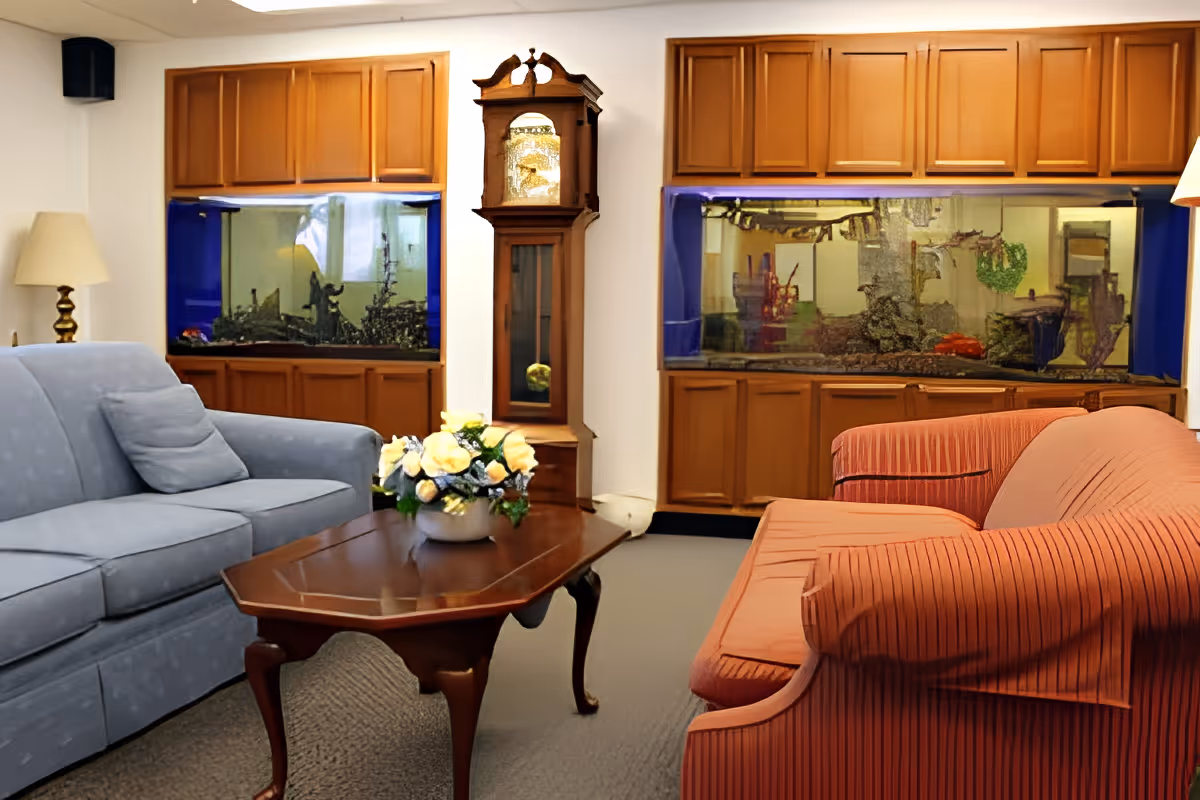 A cozy living room with a blue sofa on the left and a red striped sofa on the right, separated by a wooden coffee table with a flower arrangement. Behind the sofas are two large built-in aquariums with wooden cabinets above and below them. A tall grandfather clock stands between the aquariums, and there are lamps on either side of the room providing warm lighting.
