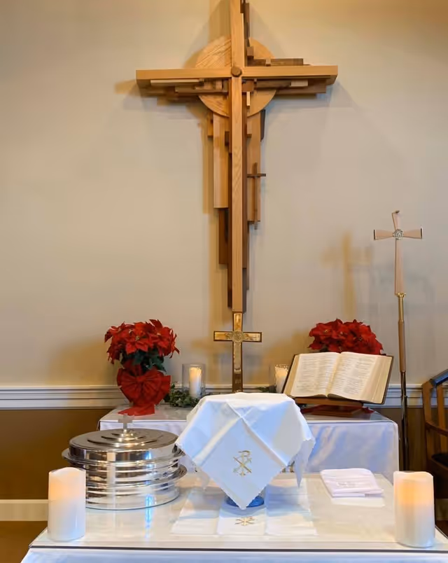 A religious altar setup inside a room with a large wooden cross mounted on the wall. The altar table is covered with a white cloth featuring a gold embroidered symbol. On the table are silver communion trays, an open Bible, two red poinsettia plants, candles, and a small cross. The background wall is beige with white trim.