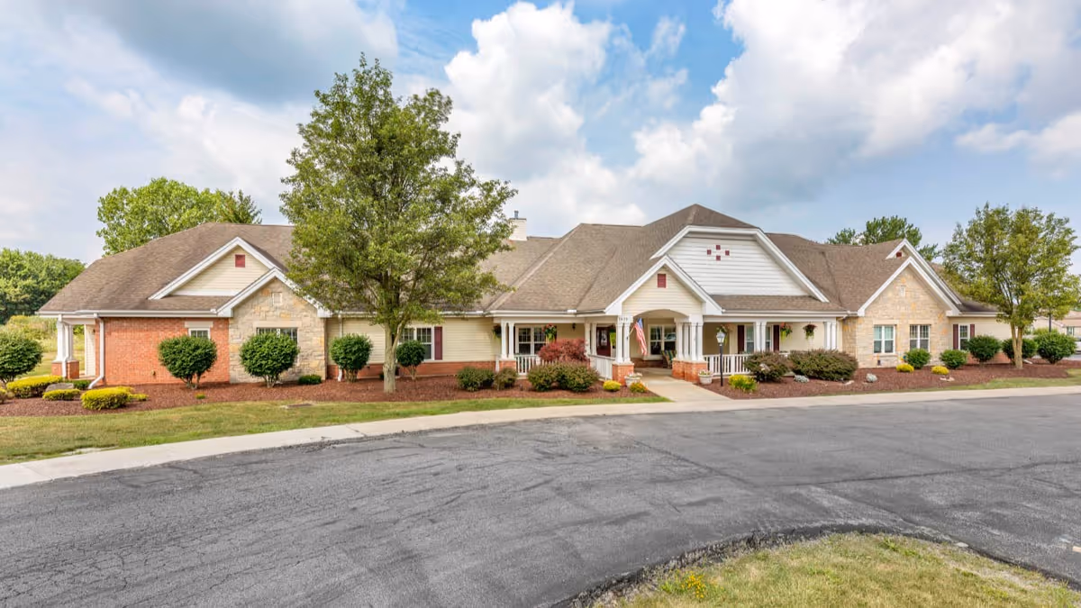 Exterior view of a single-story senior living facility building with a combination of brick, stone, and siding. The building has a covered entrance with white columns and a small porch area. There are several windows, shrubs, and trees around the building, with a paved driveway in front and a partly cloudy sky above.