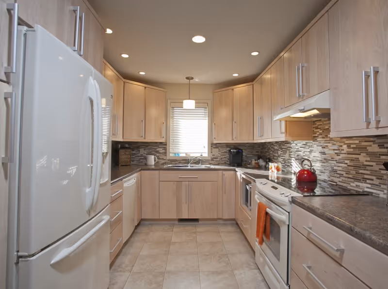A modern kitchen with light wood cabinets, a white refrigerator, a white stove with a red kettle on top, a dishwasher, and a window above the sink letting in natural light. The backsplash features a mosaic tile design in neutral tones.