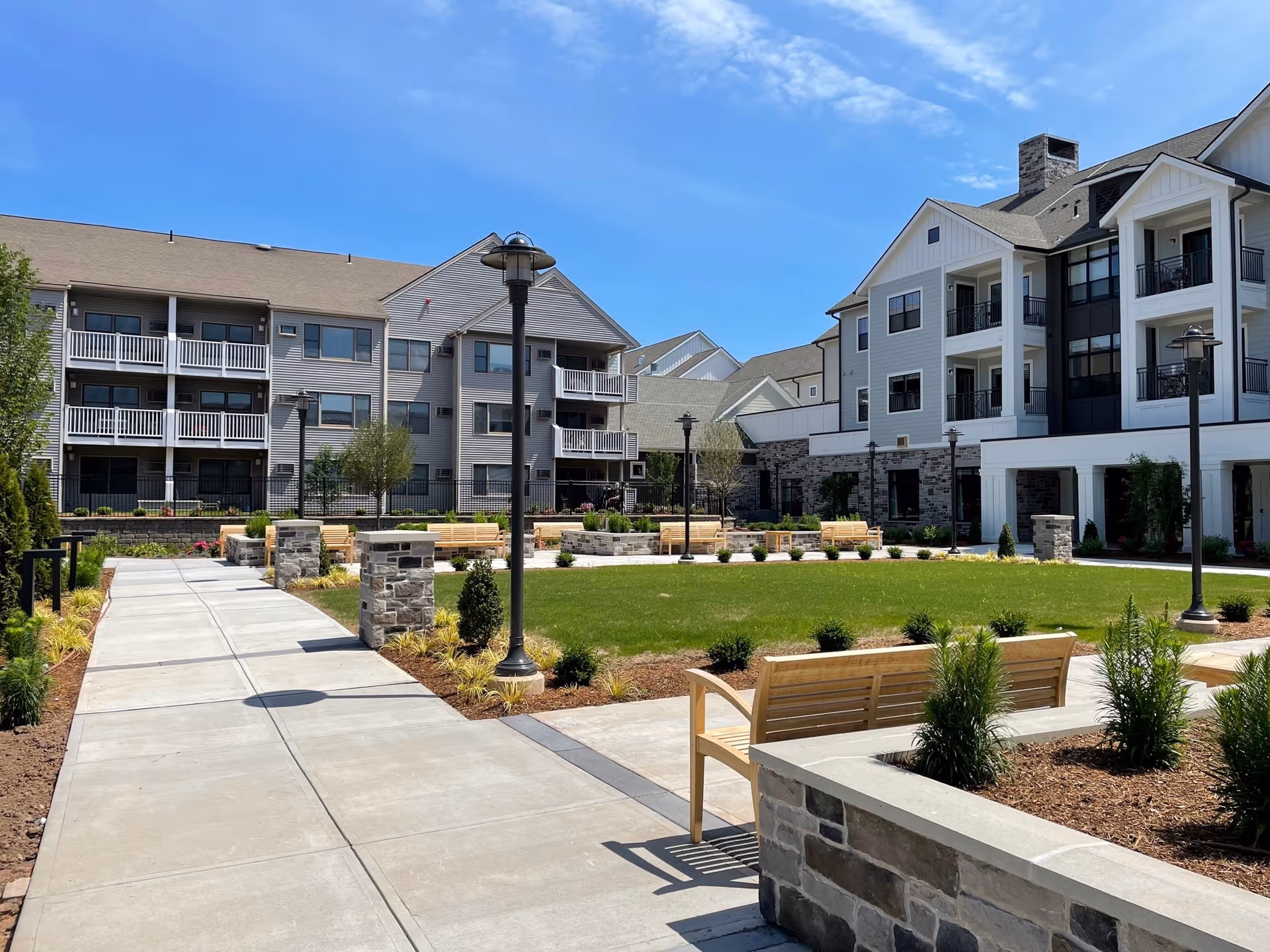 Outdoor courtyard area at Town Center at Covenant Living of Cromwell featuring a paved walkway, wooden benches, landscaped greenery, and multi-story residential buildings under a clear blue sky.