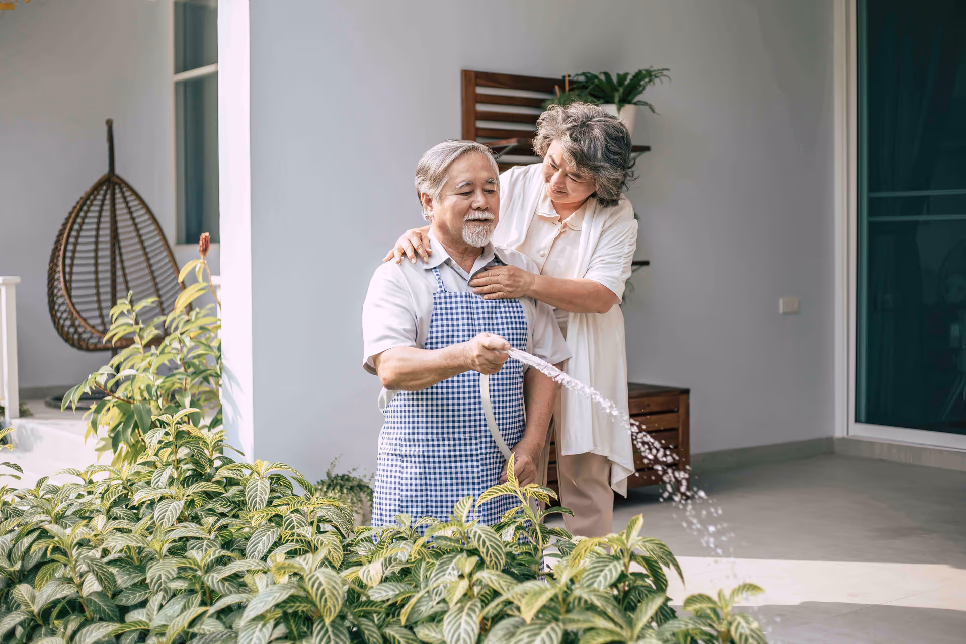 An elderly man wearing a checkered apron waters plants with a garden hose while an elderly woman stands behind him, gently placing her hands on his shoulders. They are in a bright outdoor patio area with green plants and a hanging chair in the background.