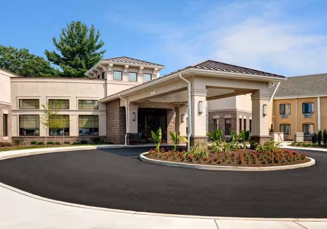 Exterior view of a senior living facility entrance with a covered drop-off area, landscaped roundabout with plants, and multiple building sections under a clear blue sky.