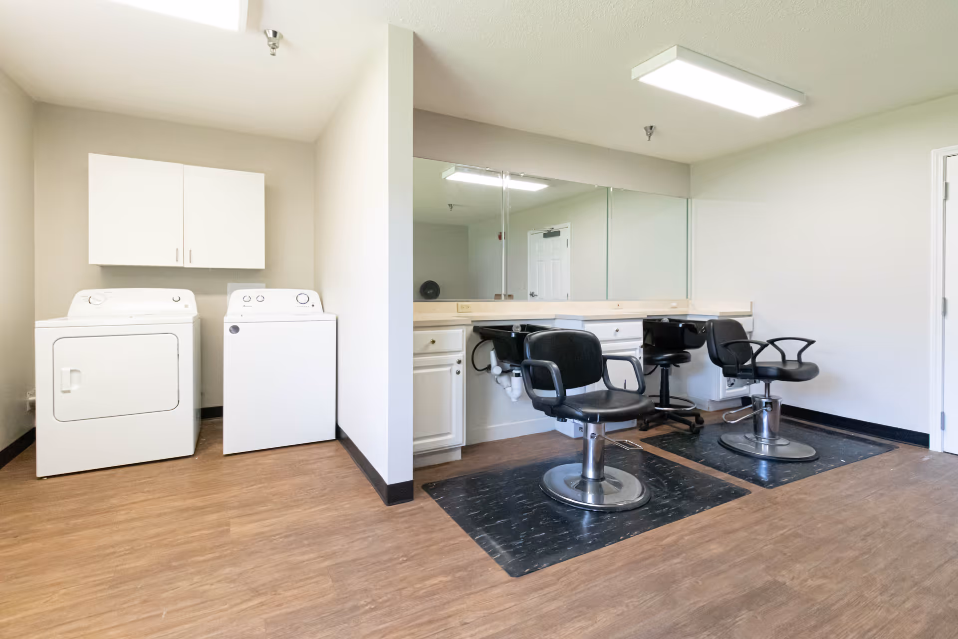 A room with two black salon chairs in front of a long mirror and countertop with white cabinets underneath. To the left, there is a white washing machine and dryer with a white cabinet mounted on the wall above them. The floor is wood laminate and the walls are painted light beige.
