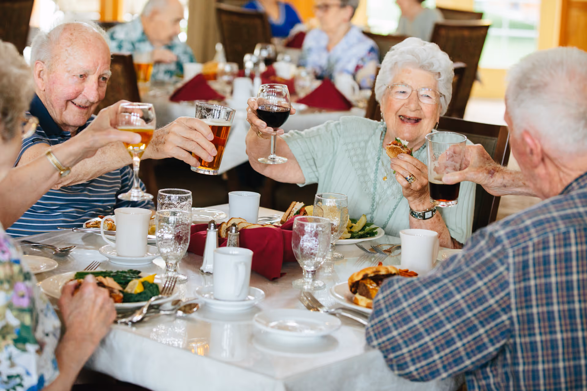 A group of elderly residents smiling and toasting drinks around a dining table.