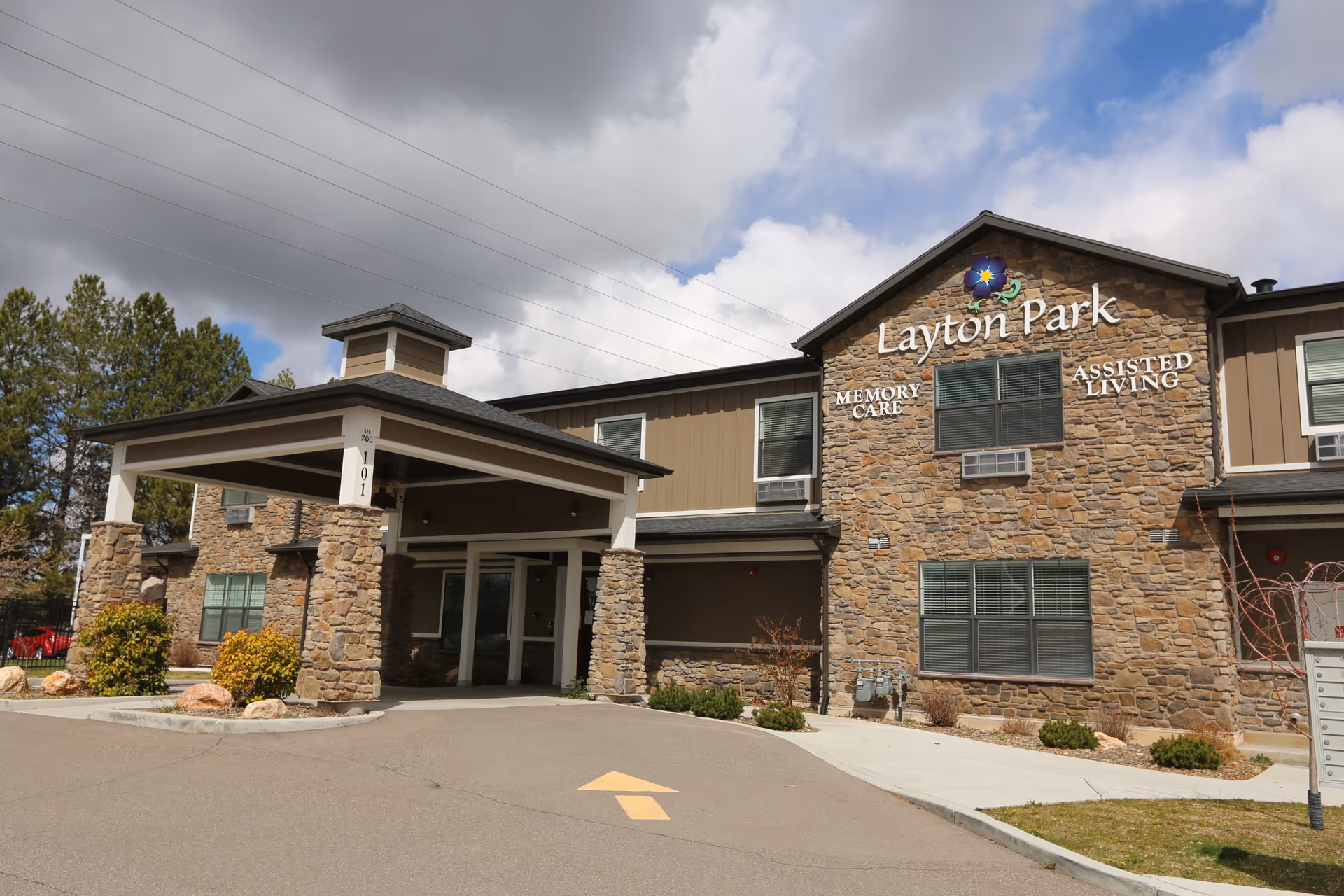 Stone-clad two-story assisted living and memory care building with a covered entrance and a driveway arrow.
