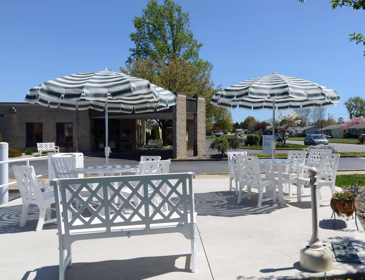 Outdoor patio area with white chairs and tables under green and white striped umbrellas, situated in front of a building with trees and a clear blue sky in the background.