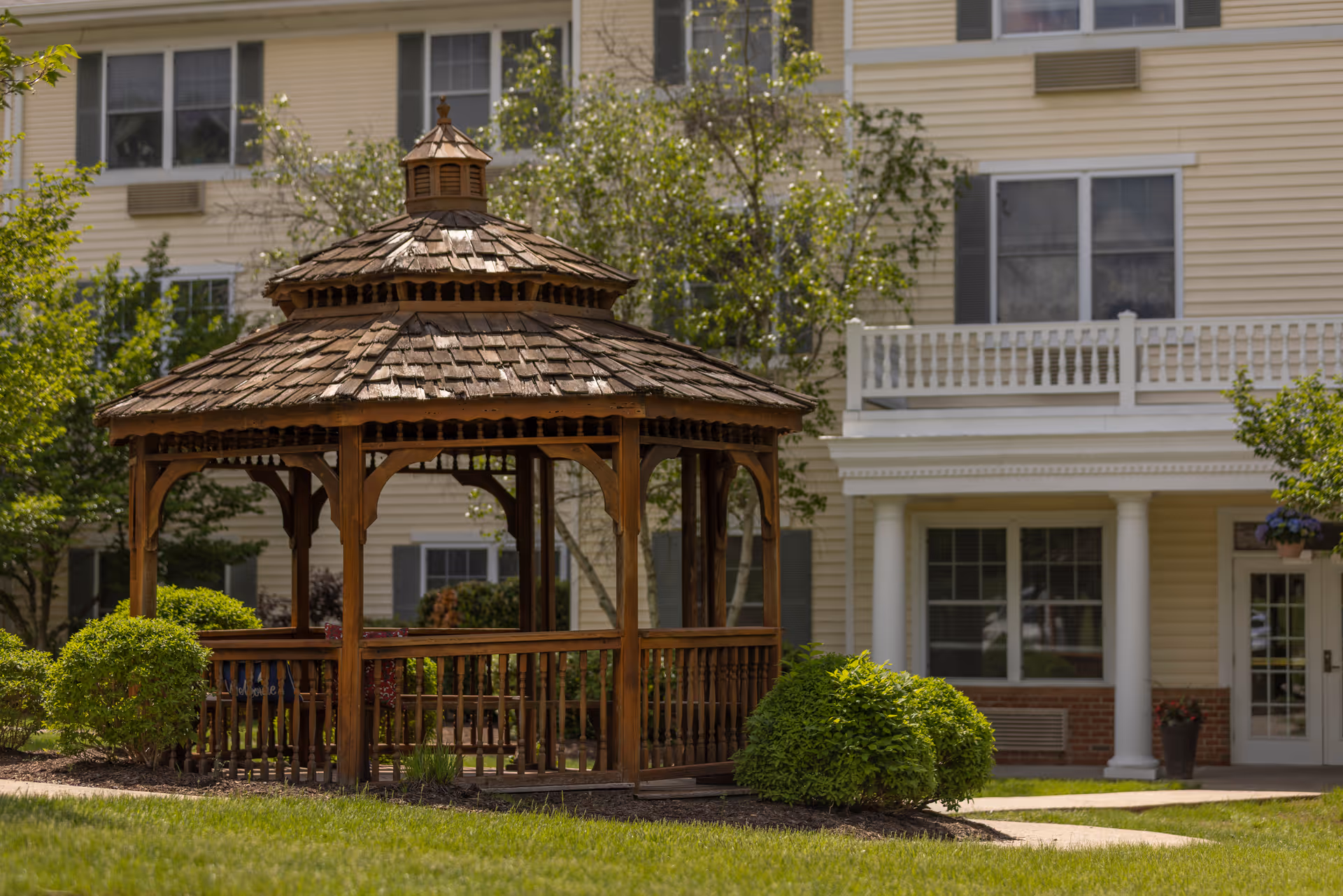 A wooden gazebo with a shingled roof situated on a grassy area with bushes and trees, in front of a beige multi-story building with white trim and windows.