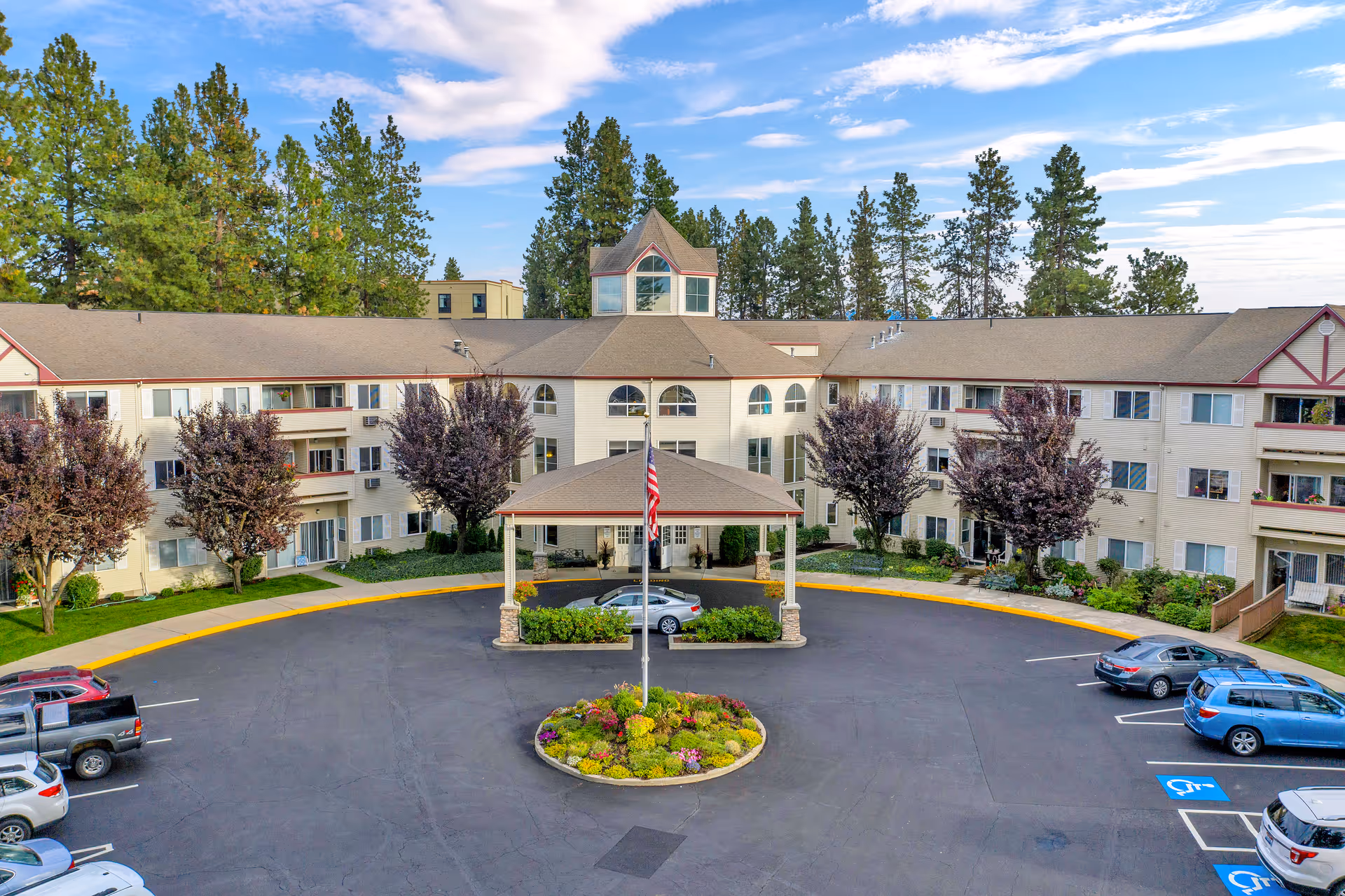 Front exterior of a three-story senior living facility with a covered circular driveway, landscaped center island, and parked cars.