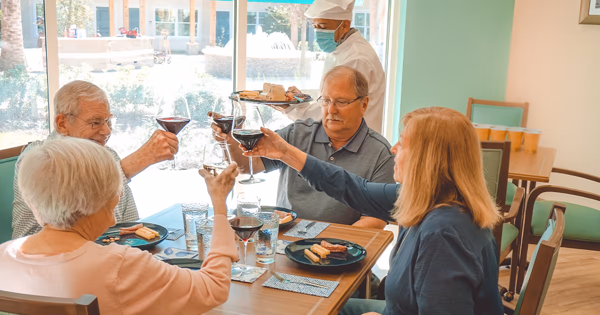 Four older adults seated at a dining table raise wine glasses in a toast while a masked server stands behind them with a tray of food.