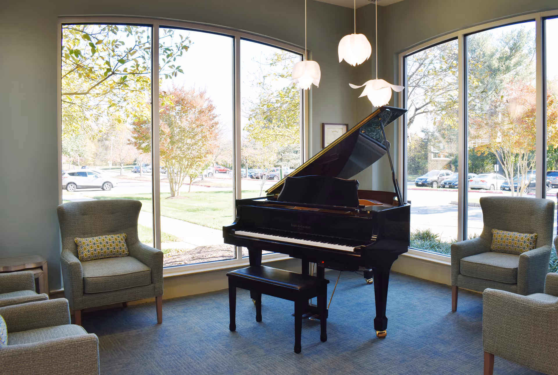 Sunlit lounge with a black grand piano centered in front of large arched windows and upholstered armchairs.