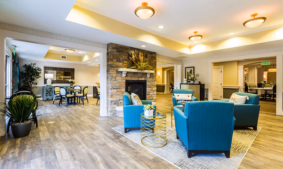 A bright and spacious senior living common area featuring a stone fireplace surrounded by four teal armchairs arranged around a small round gold and glass table. The room has wood flooring, recessed lighting, and a dining area with a table and chairs visible in the background. Potted plants and decorative elements add warmth to the space.