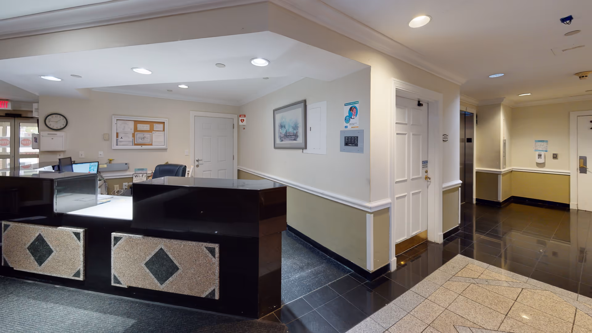 Reception area of a senior living facility with a black and beige front desk, a clock on the wall, a bulletin board, a framed picture, and doors leading to other rooms and an elevator.