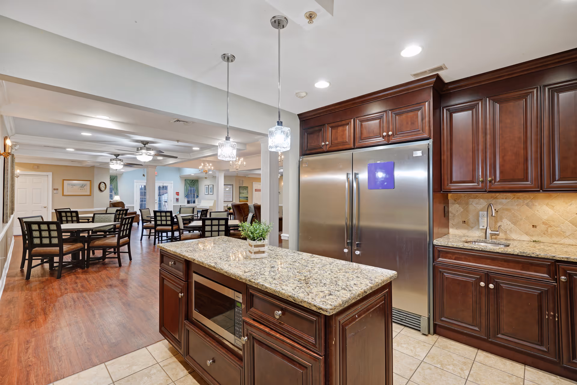 Interior view of a senior living facility kitchen and dining area. The kitchen features dark wood cabinets, a large stainless steel refrigerator, a granite countertop island with a built-in microwave, and a small plant on the island. The dining area has several tables and chairs with wooden floors and ceiling fans, extending into a well-lit common area.